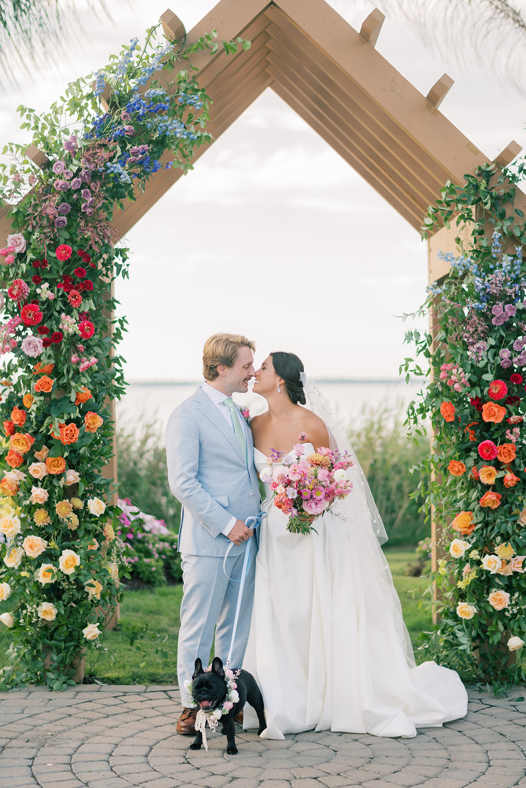 bride and groom pose with dog under wooden arbor with colorful roses 