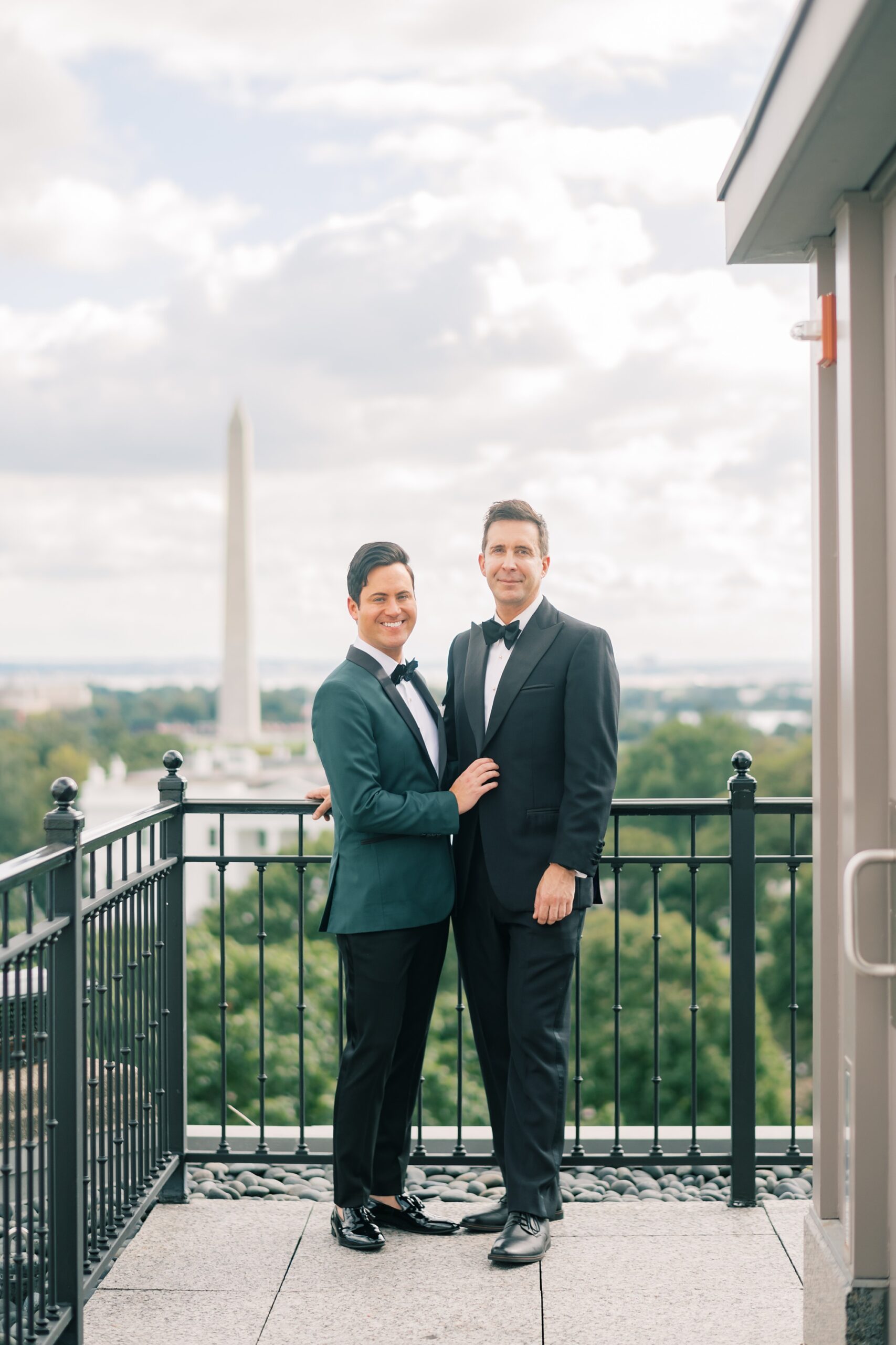 two grooms stand on balcony of Hay Adams Hotel in Washington DC on wedding morning overlooking city