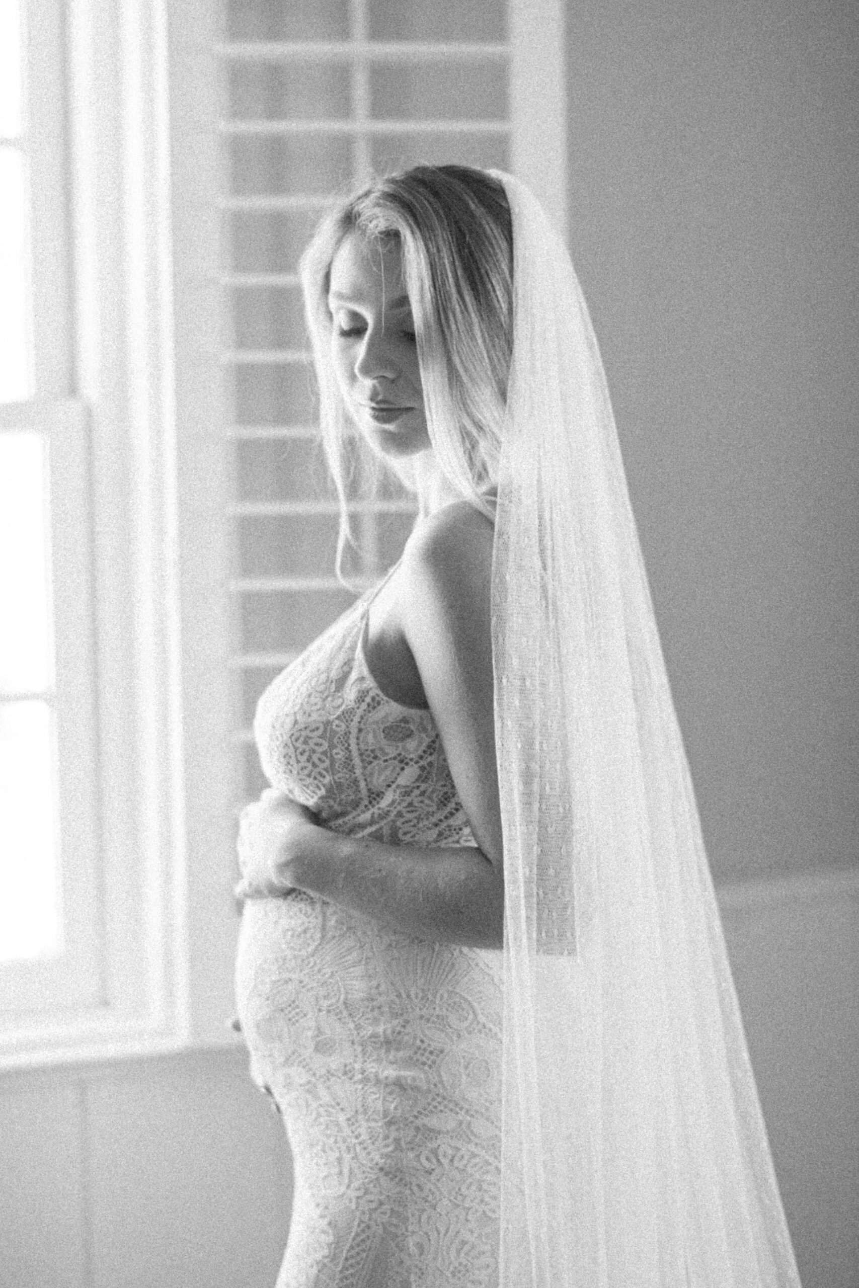 pregnant bride stands holding baby bump with veil around shoulders 