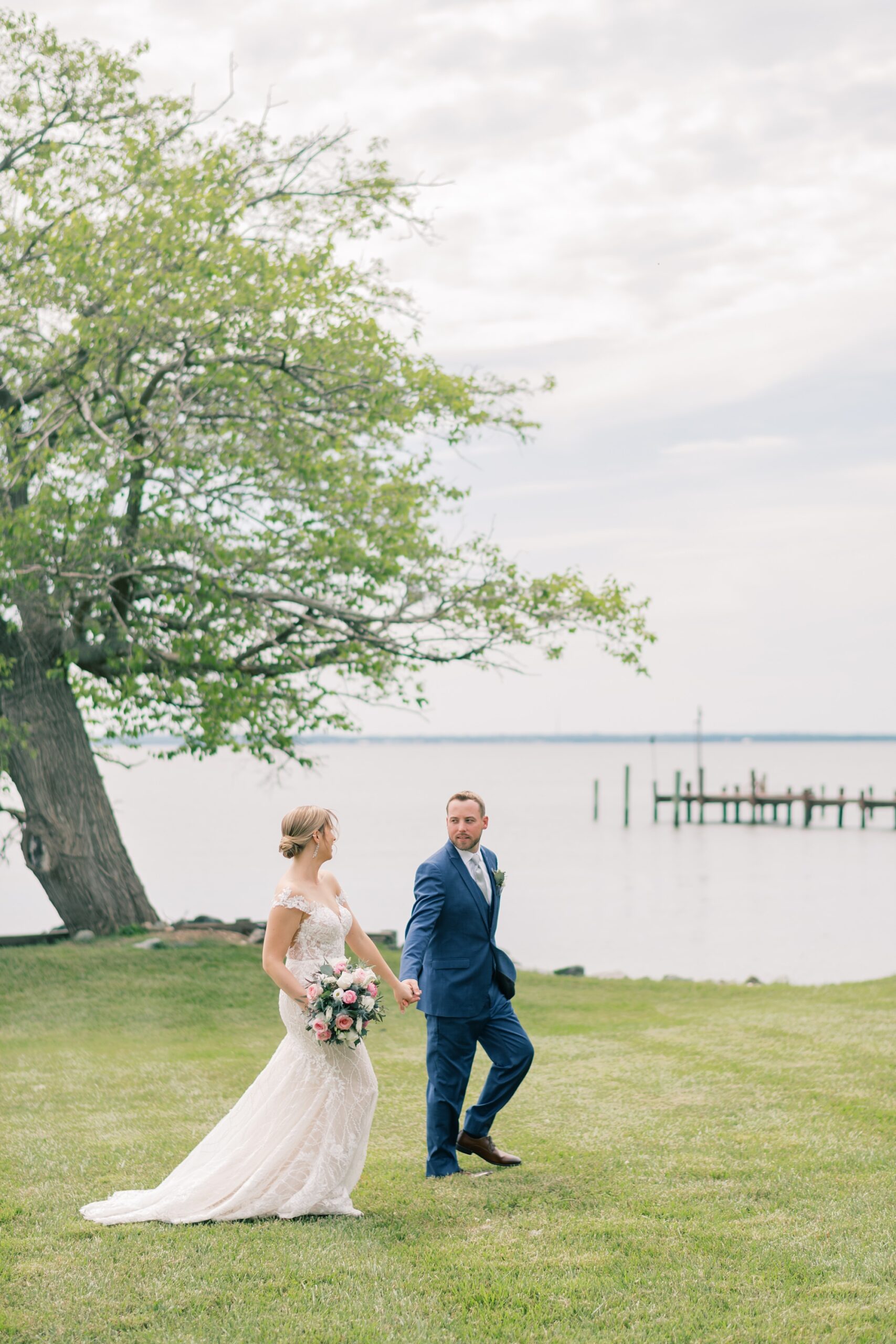newlyweds hold hands walking across lawn with water behind them at the Pavilion at Weatherly