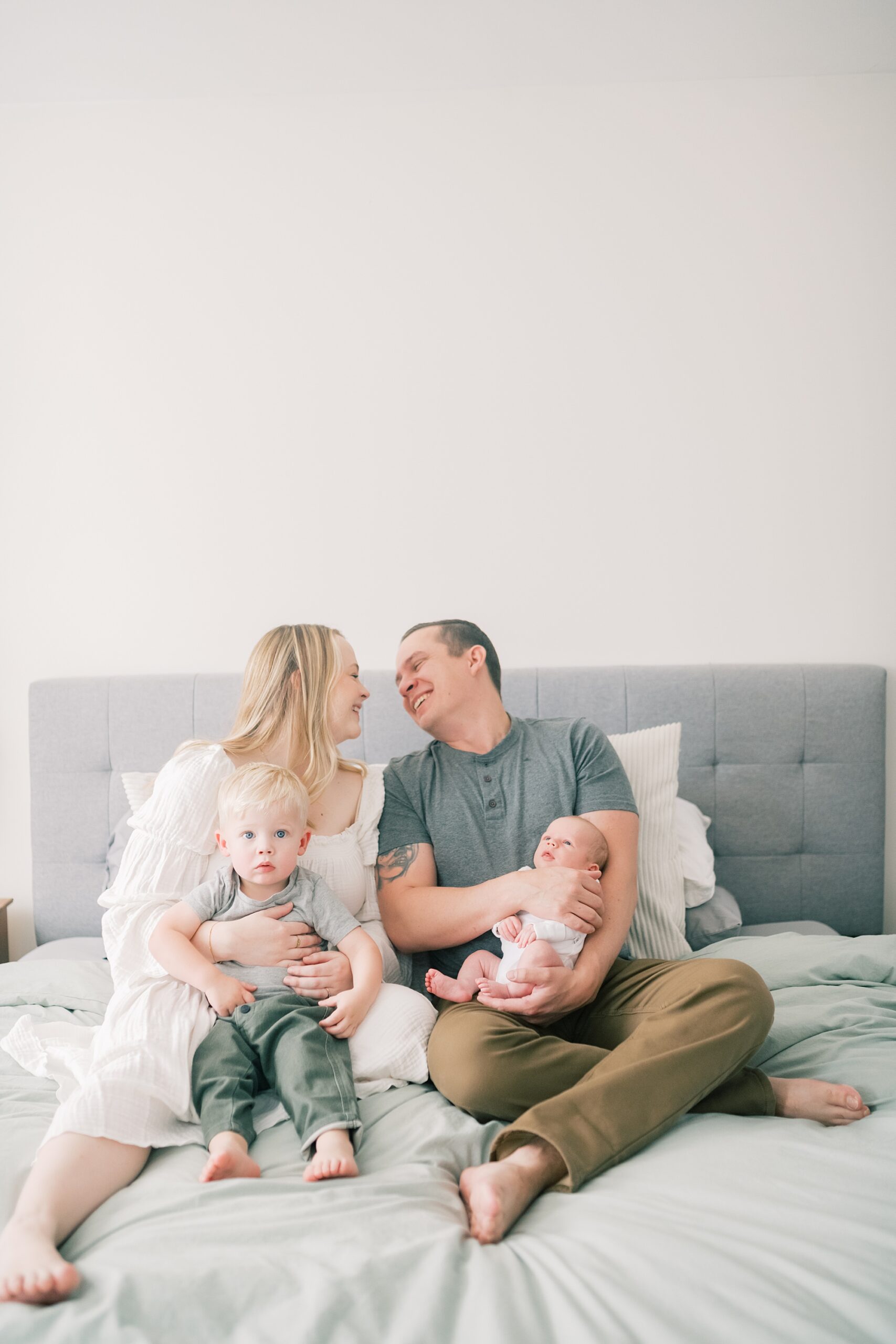 couple laughs holding two sons on bed during in-home newborn portraits