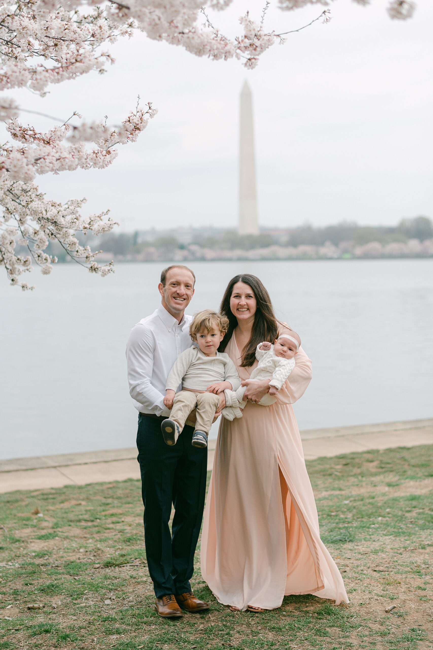 family of four poses under cherry blossoms in front of Washington Memorial during Cherry Blossom Portraits at the Tidal Basin