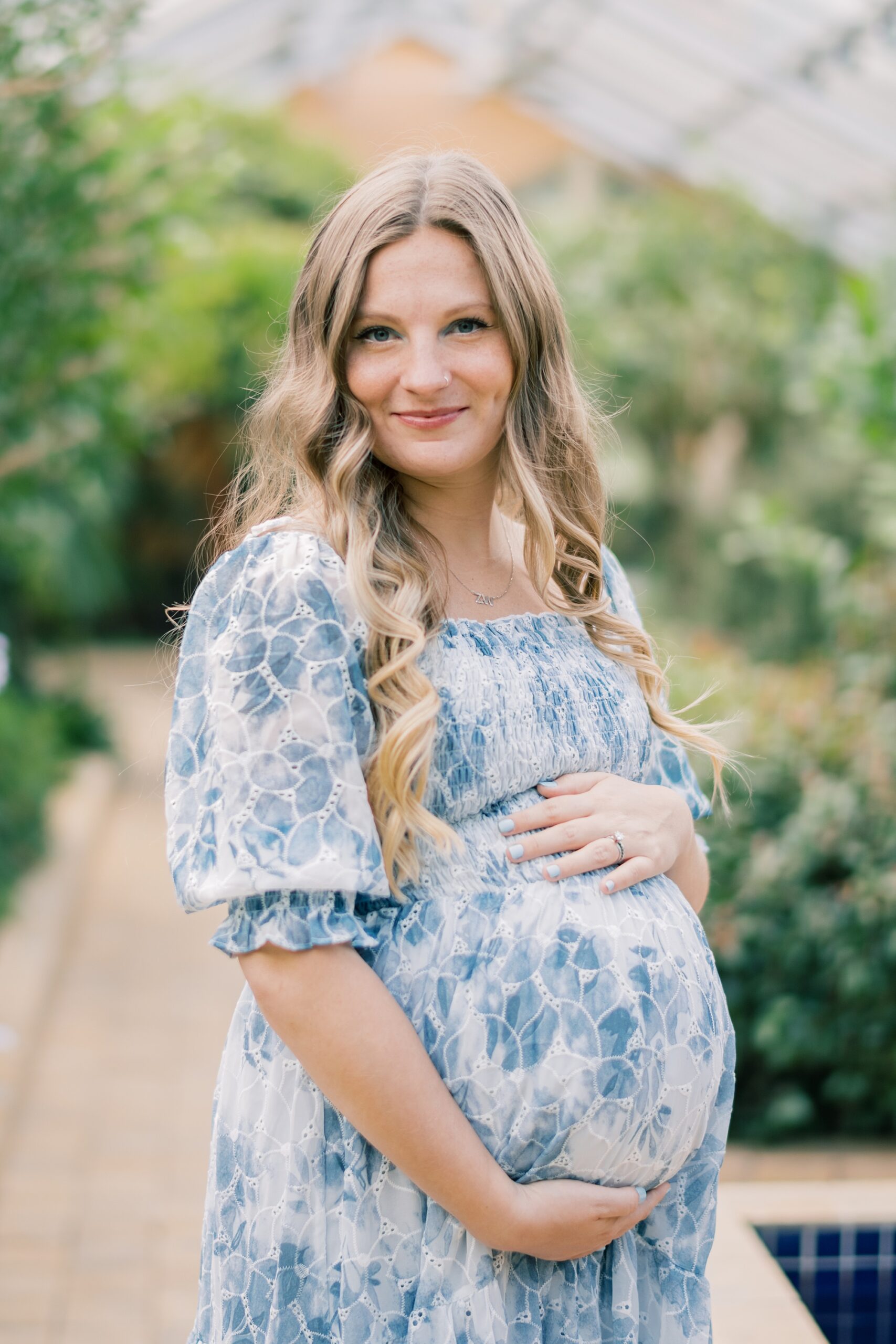 woman smiles holding baby bump in blue and white dress at Rawlings Conservatory