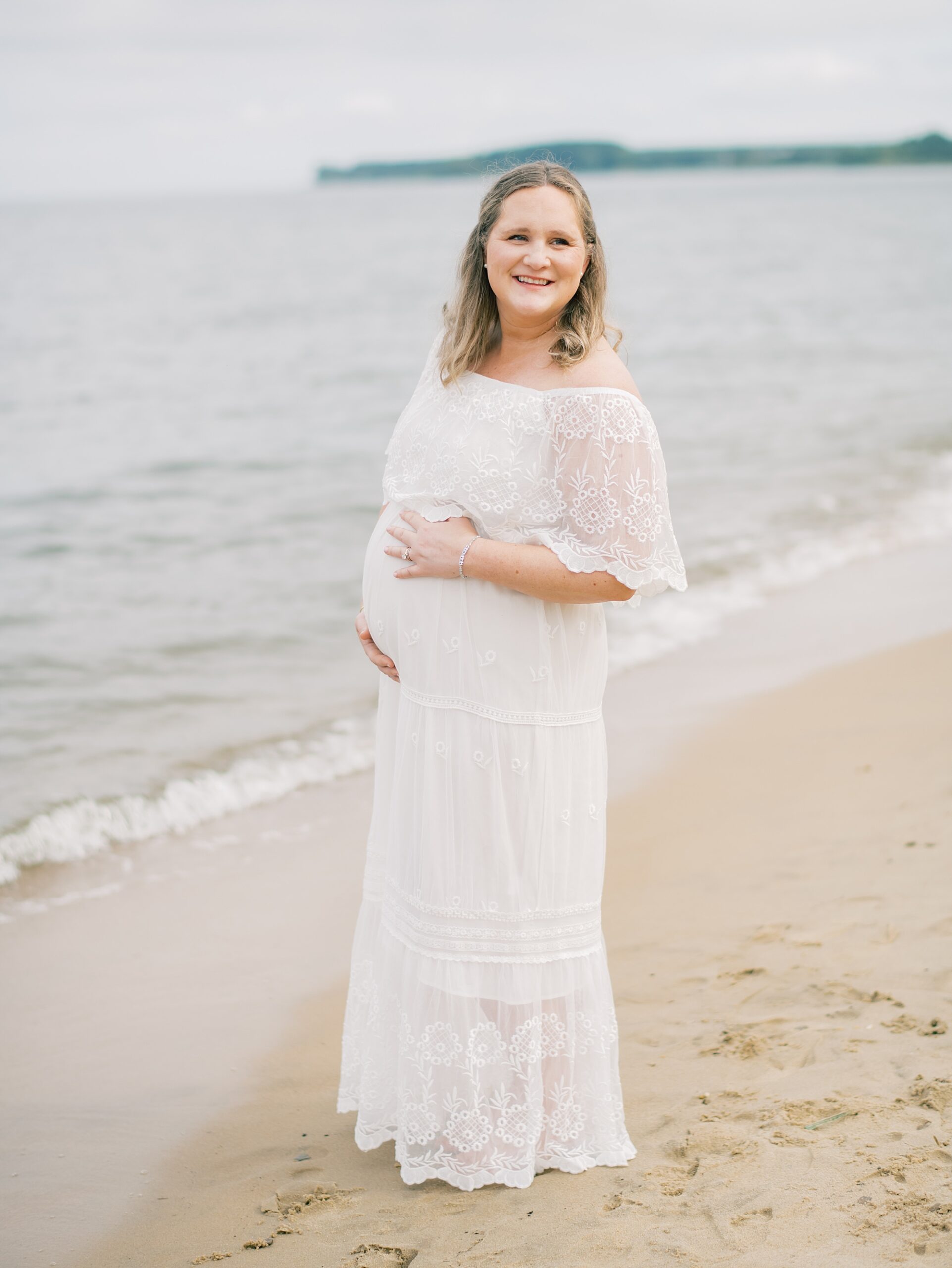 expecting mother holds baby bump standing on the beach during maternity session in Annapolis Maryland