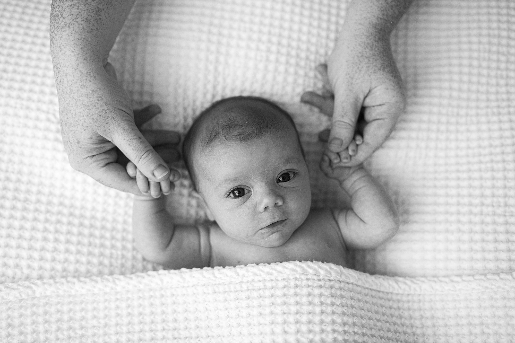 baby girl lays on white sheet holding mom's hands during studio newborn session in Solomons Maryland