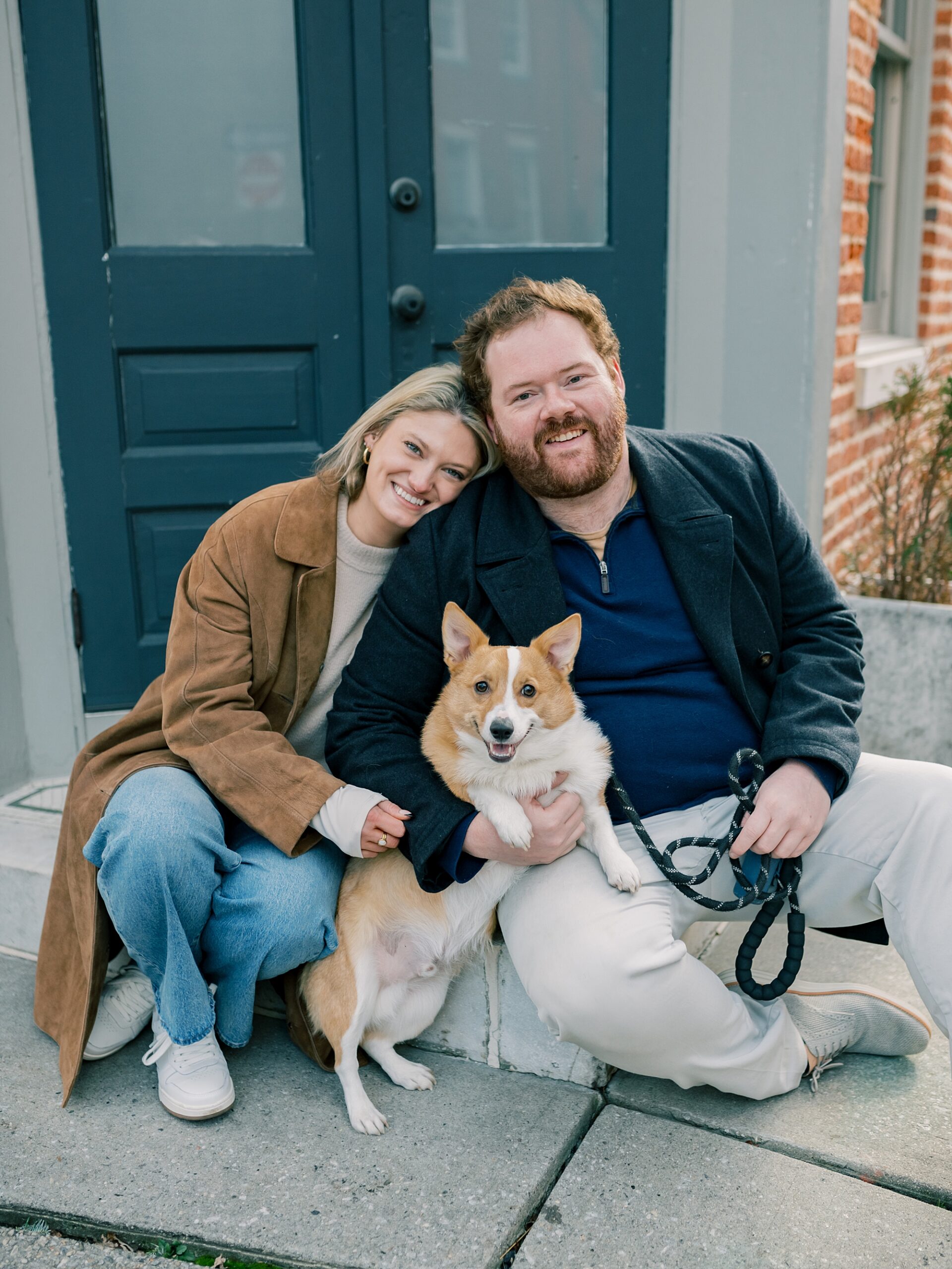 engaged couple sits with corgi on front step during Baltimore engagement session