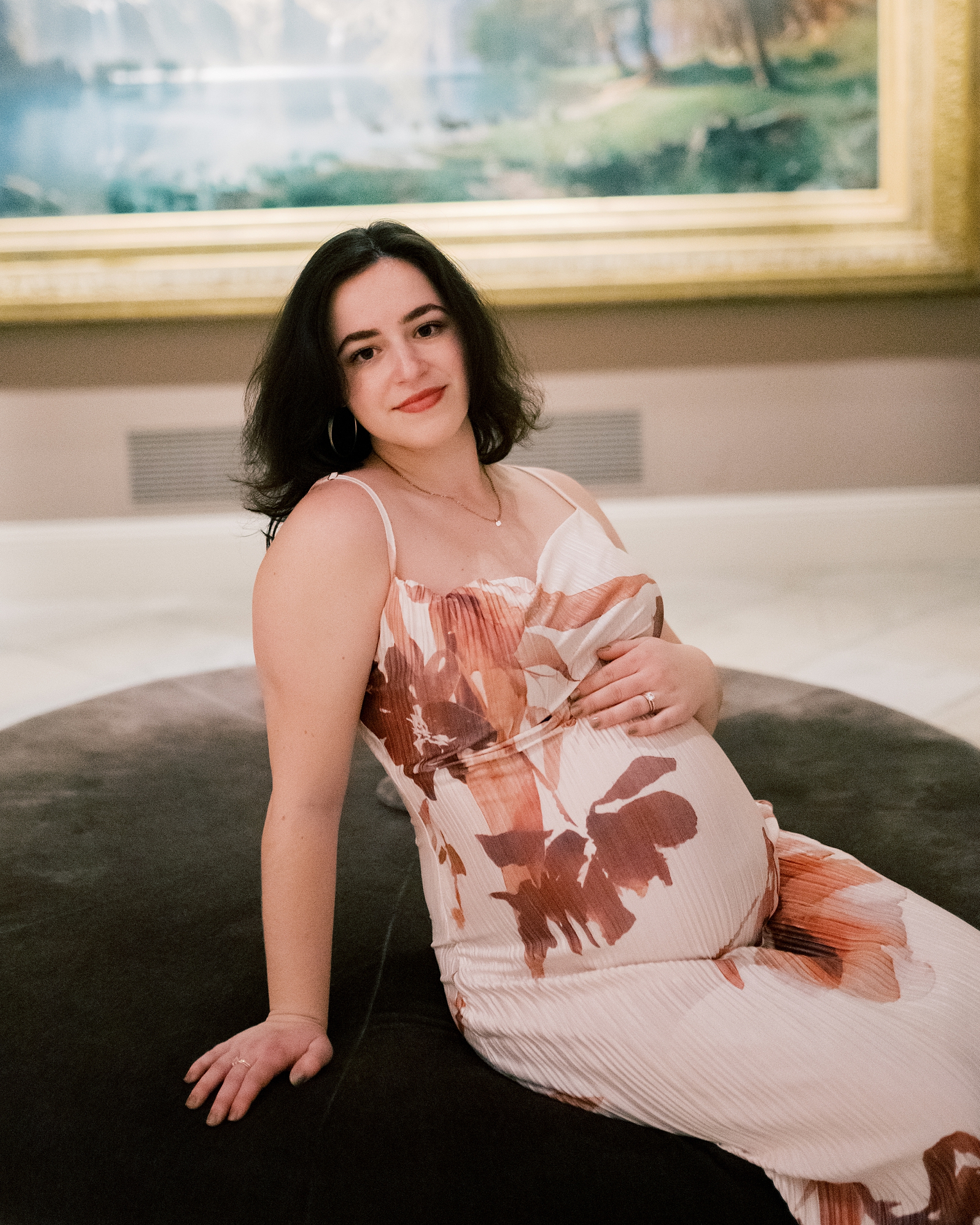 woman sits on tan ottoman holding baby bump during maternity session at the National Portrait Gallery