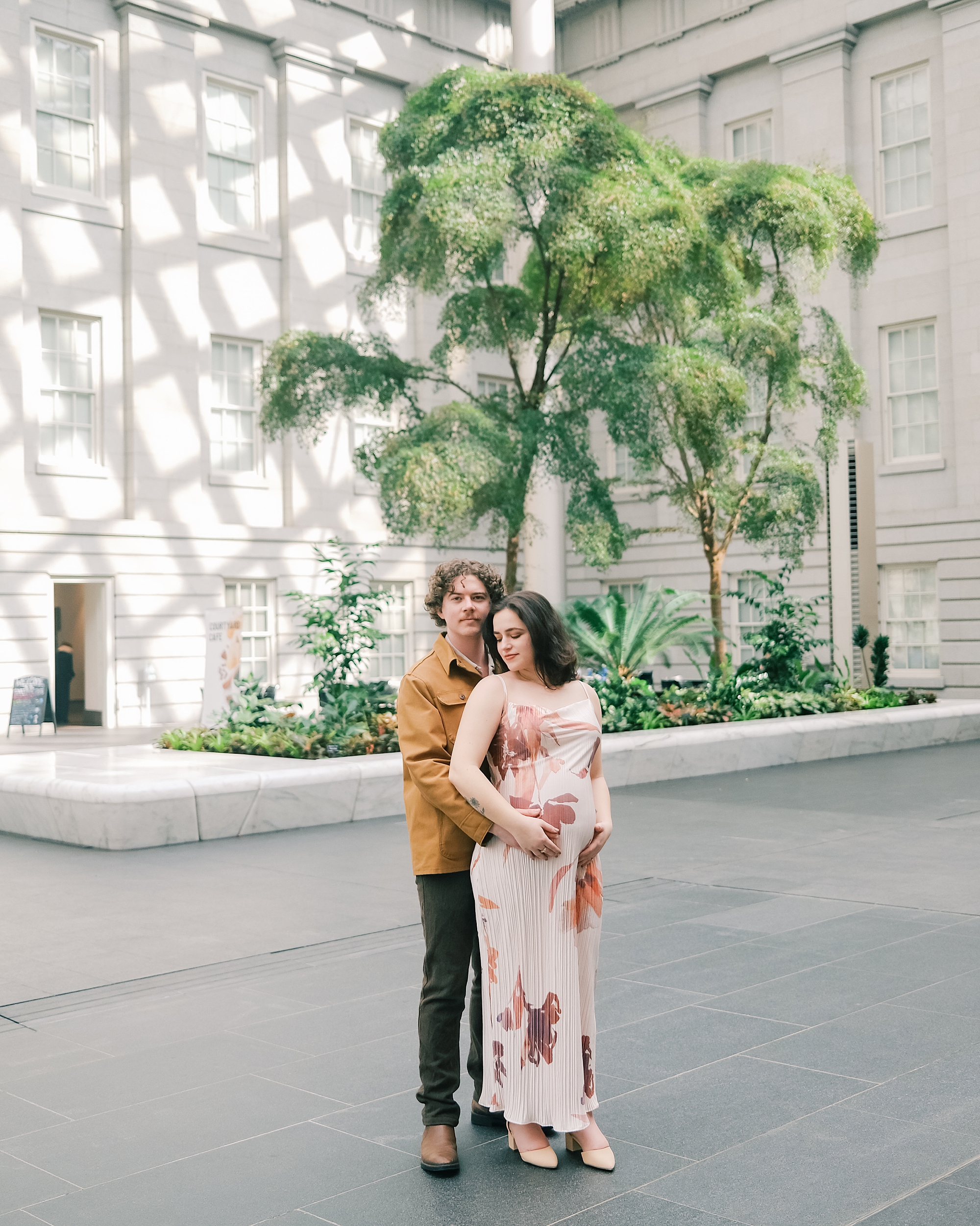 man stands behind woman holding her baby bump during maternity session outside the National Portrait Gallery