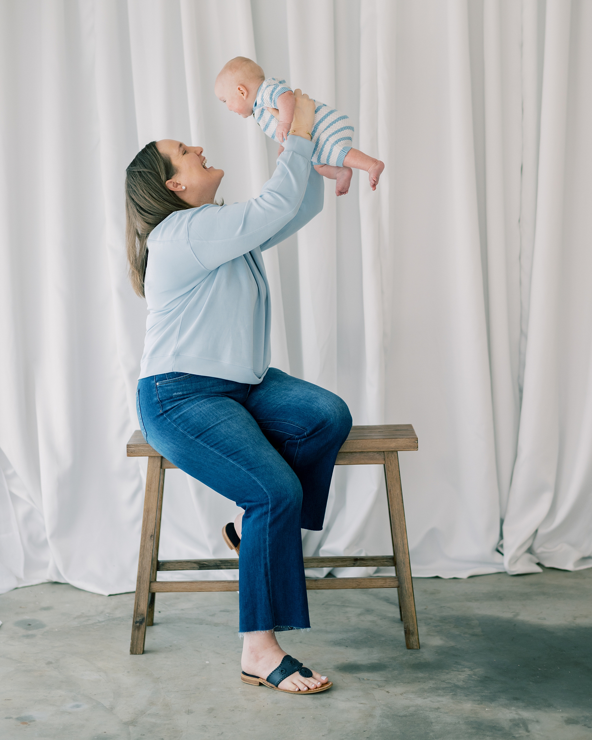 mom sits on stool holding baby boy up in the air during milestone session in Southern Maryland studio