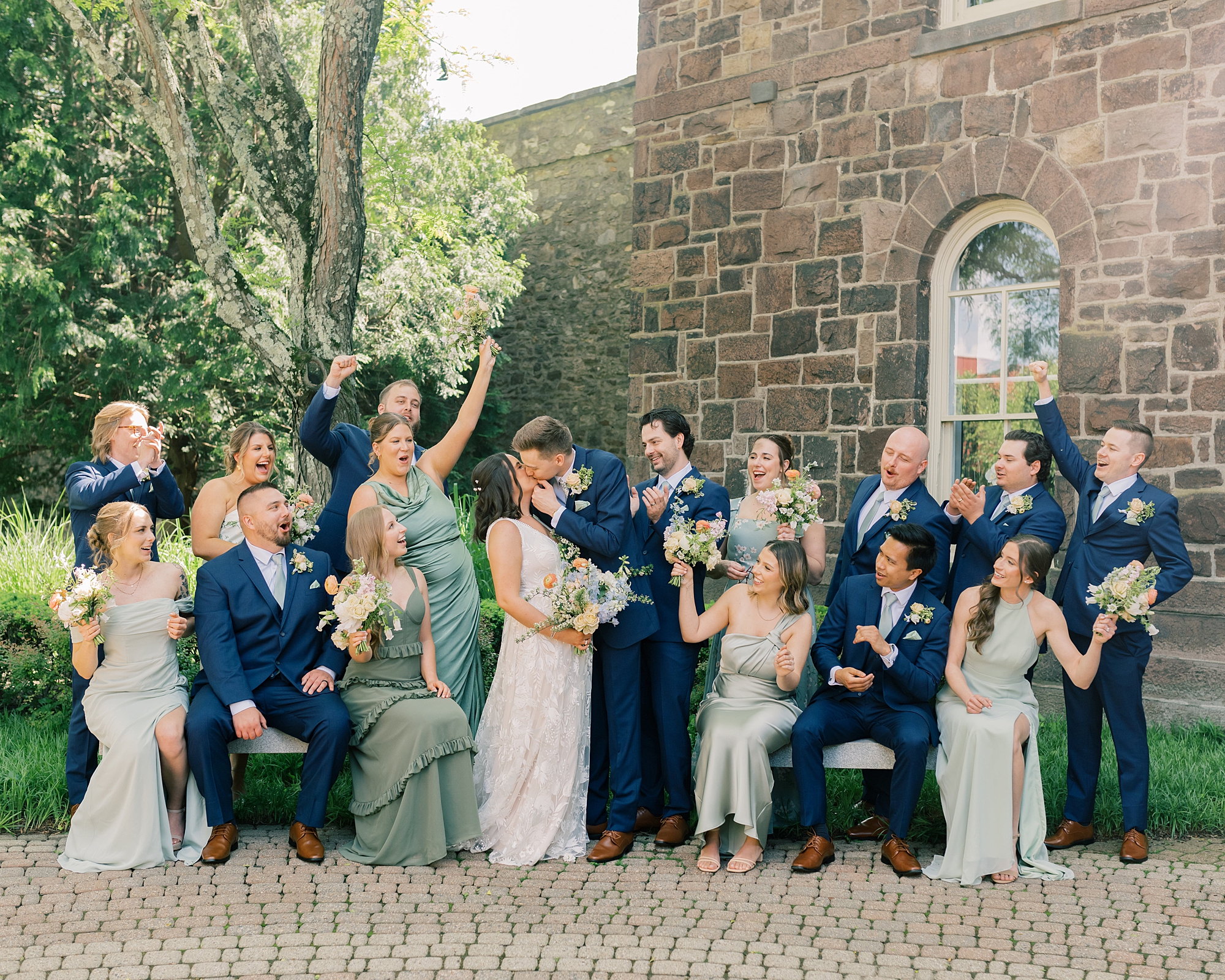 wedding party cheers outside brick building while bride and groom kiss