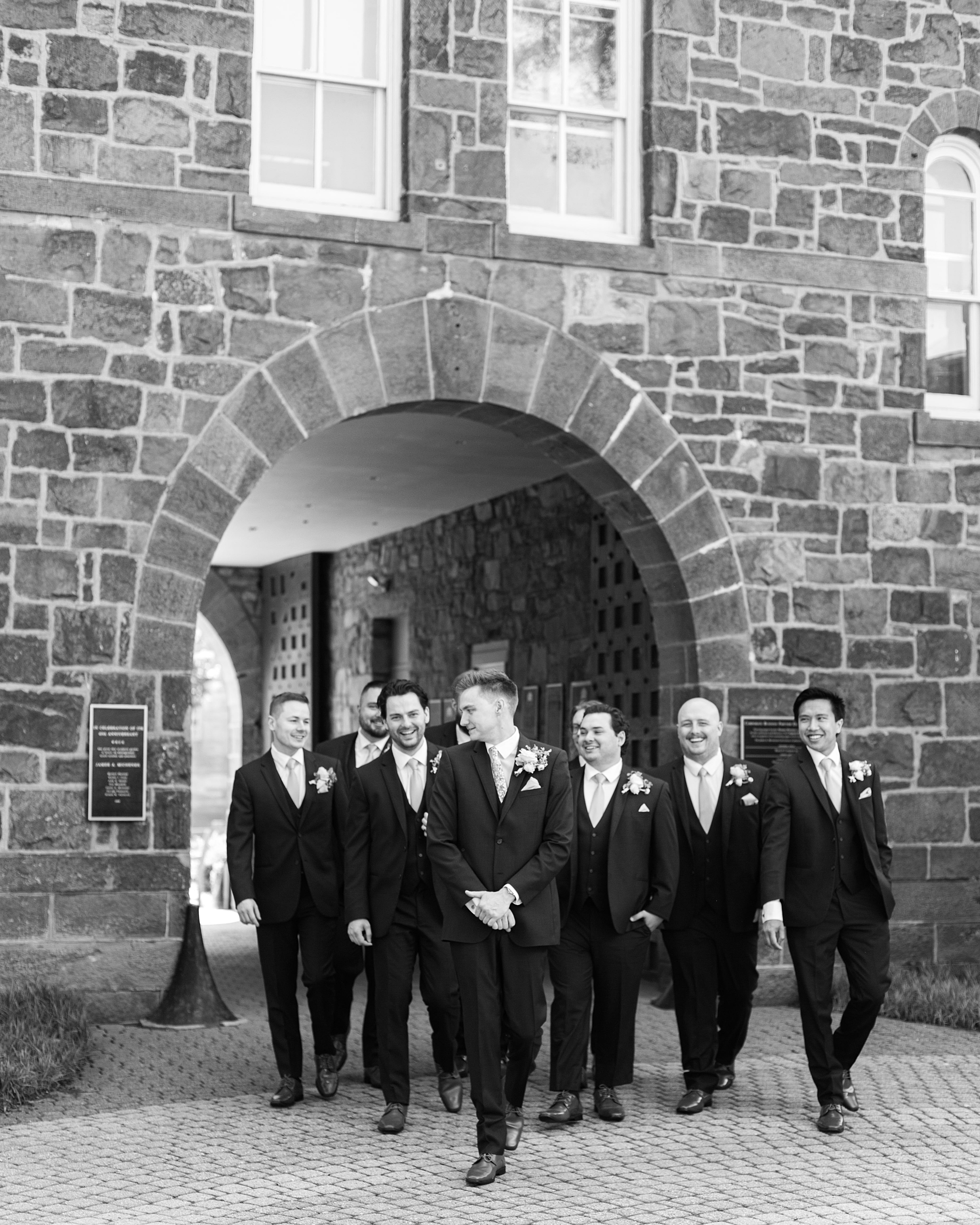 groom walks with groomsmen under arch of stone building
