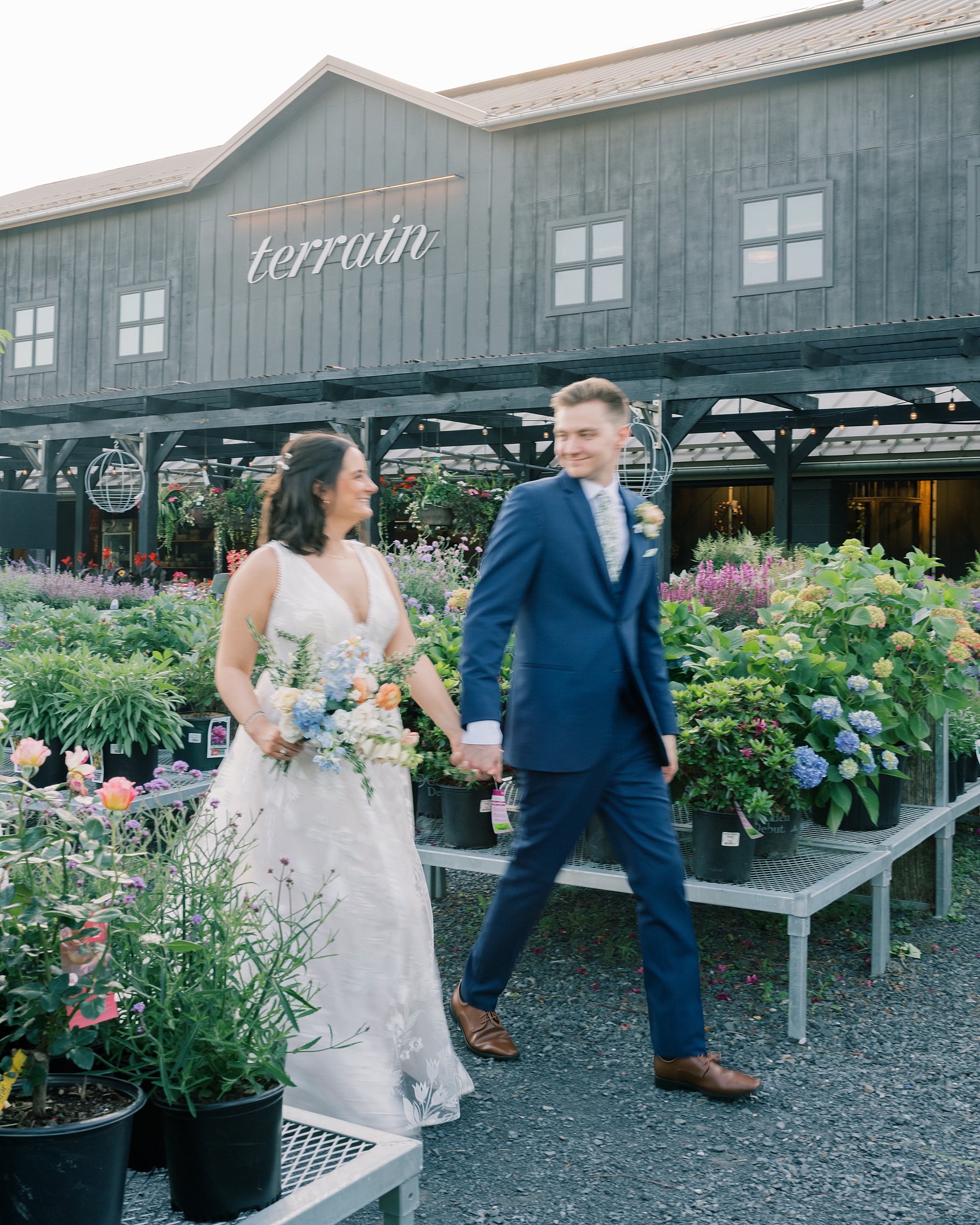 groom holds hands with bride walking between rows of flowers at Terrain Gardens