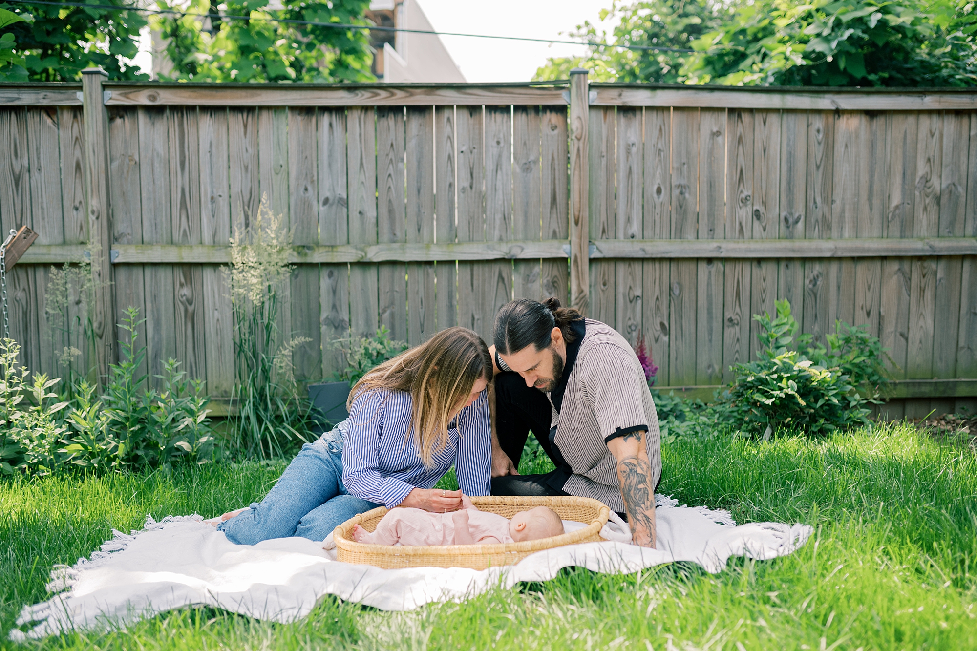 parents sit on blanket looking down at baby girl in Moses basket