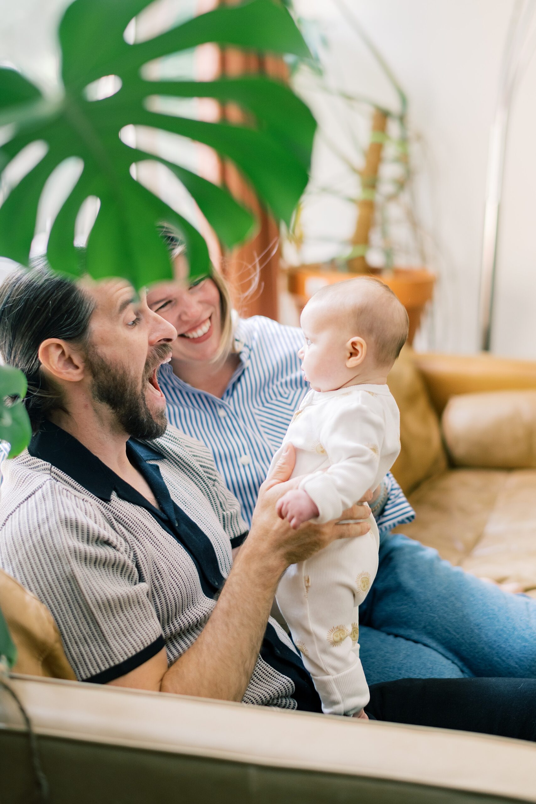 parents laugh with baby in front of them behind monstera plant