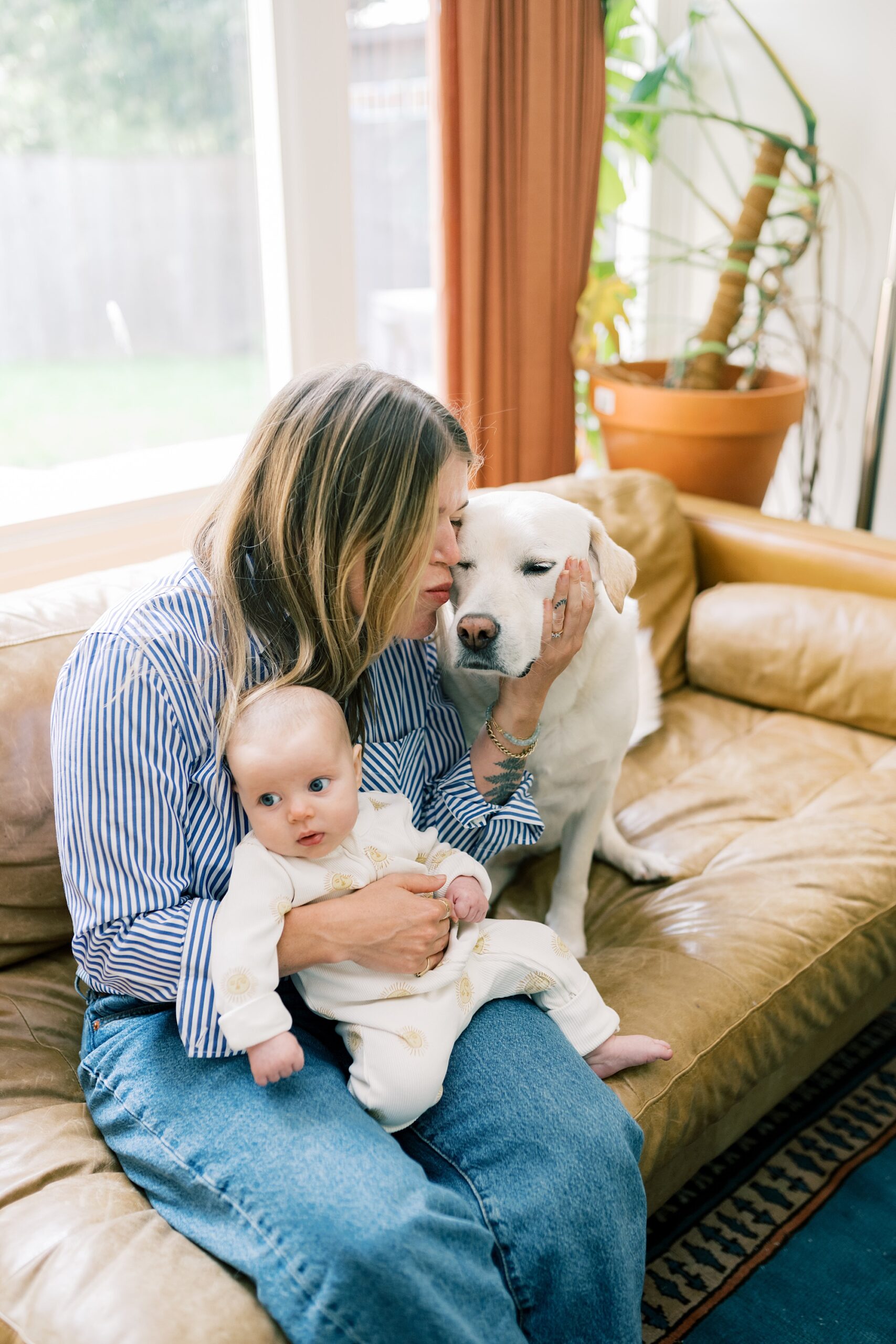 mom holds baby on lap and kisses dog on couch