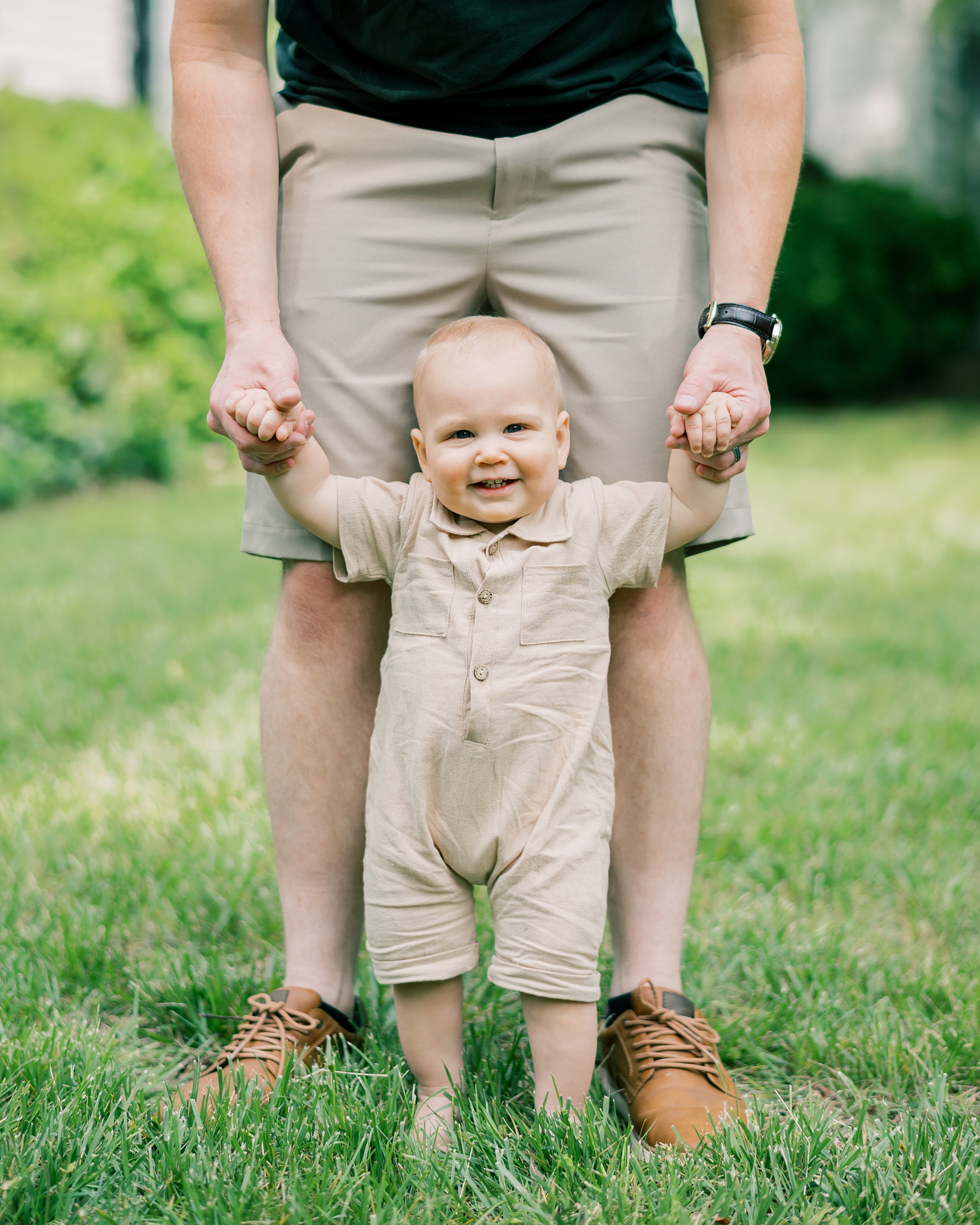 toddler in tan outfit stand on dad's toes on lawn
