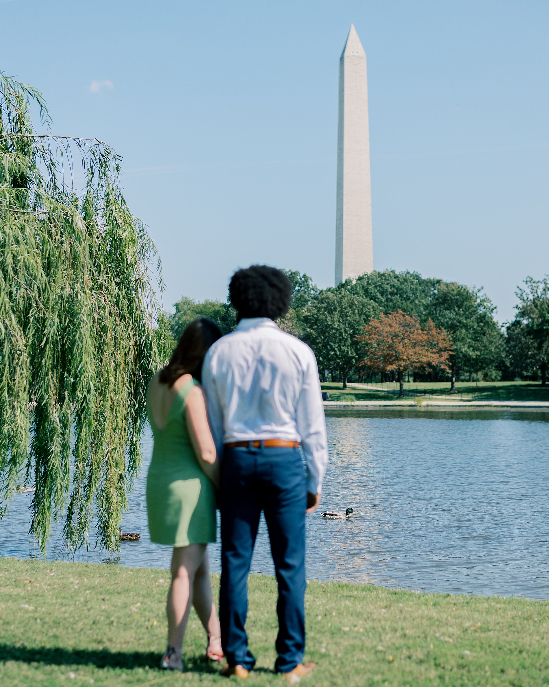 couple with their heads leaning on each other as they watch ducks in pond with the Washington monument in the distance