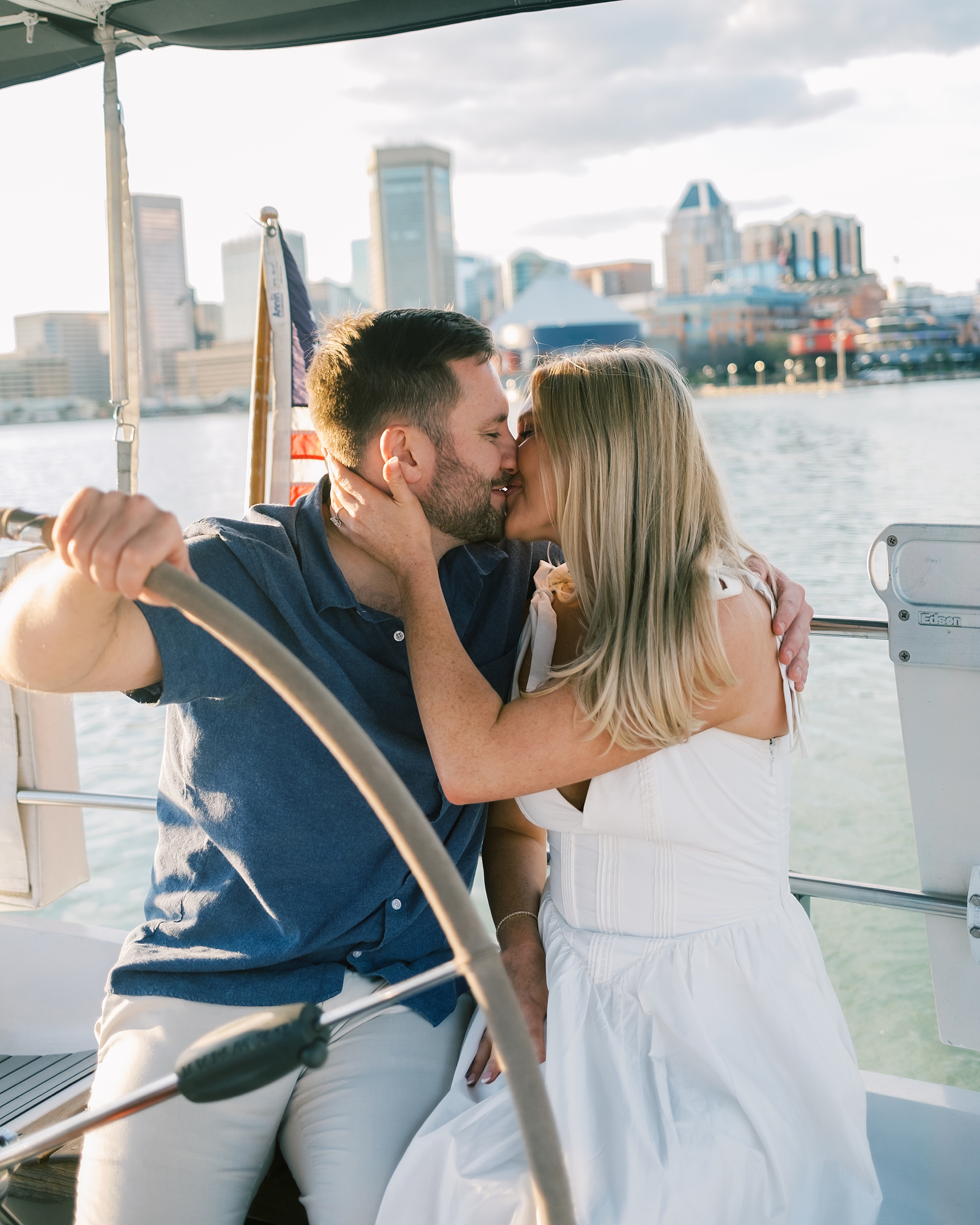 engaged couple kiss on sailboat with Baltimore skyline behind them 
