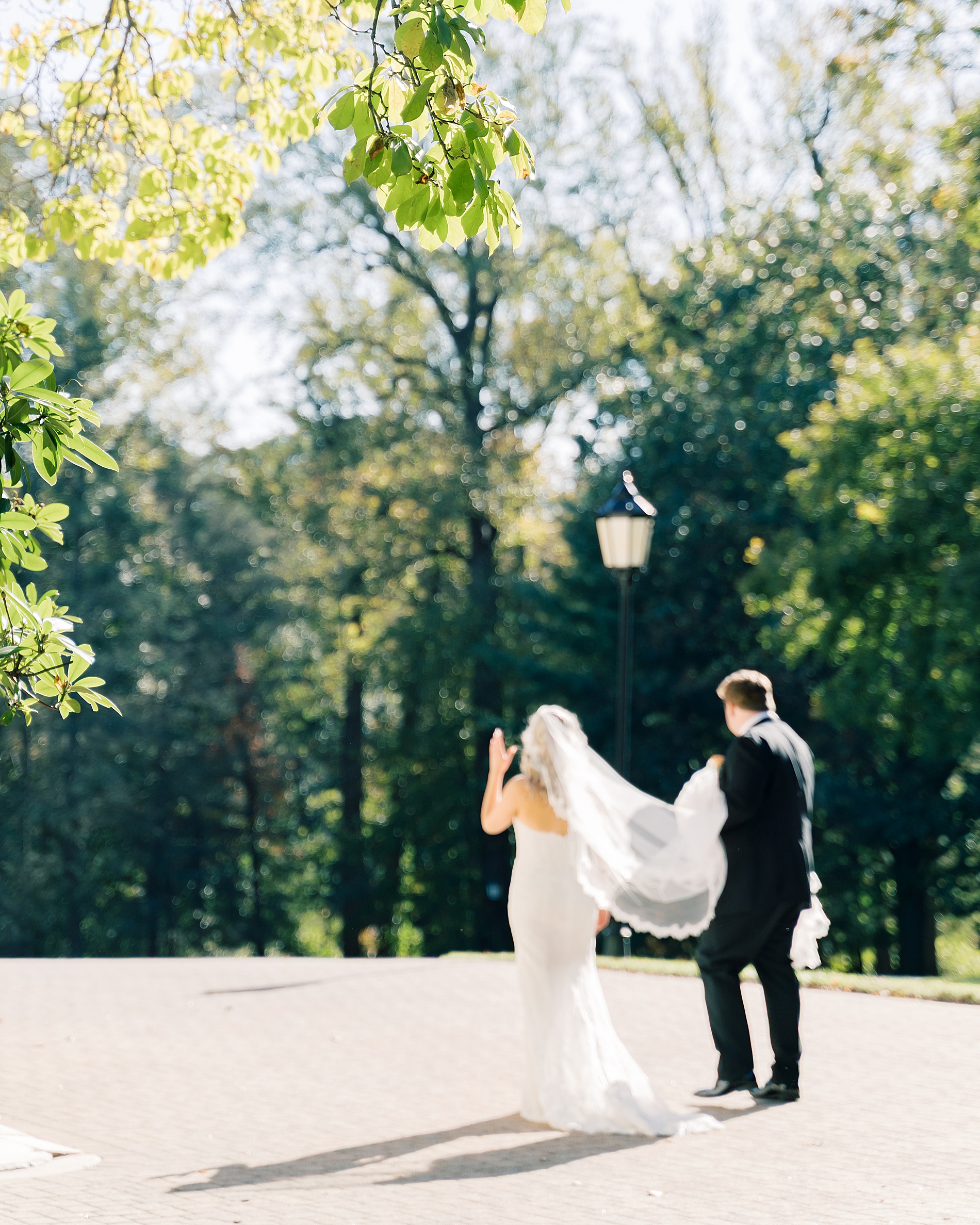 groom carries brides long veil as they walk together 