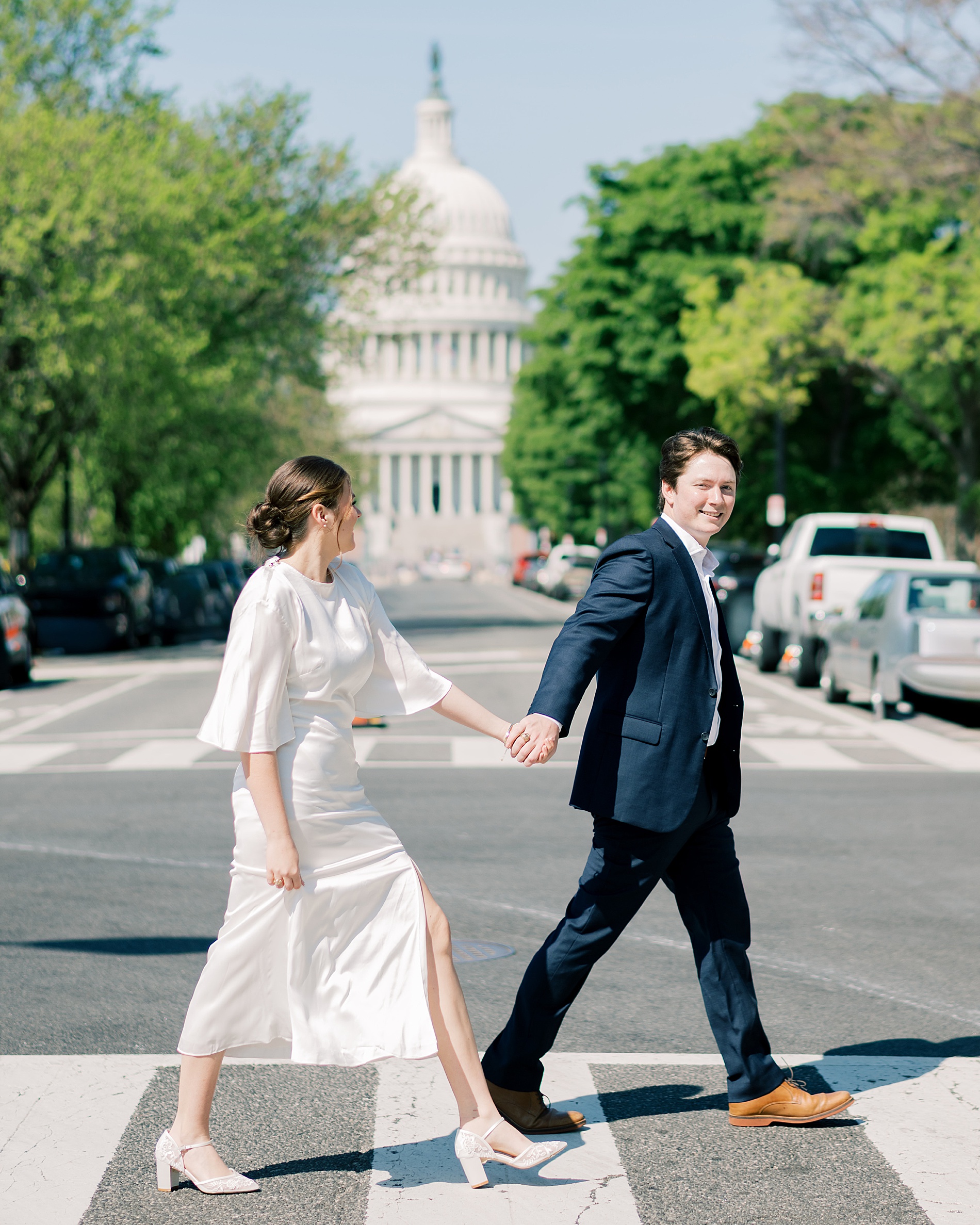 man holds his fiancé's hand as they cross street with Capitol building in background 